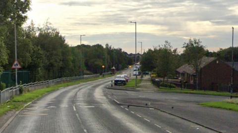 A road with speed bumps and yellow zig zag lines. In the background is parked vehicles and a zebra crossing with flashing lights on pillars. 
