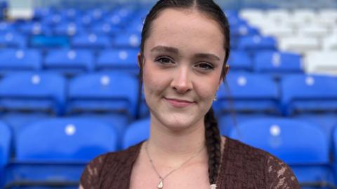 Jess McGhee, standing in Peterborough United's stadium. She has dark brown hair pulled back into a neat plait over her left shoulder and is wearing a dark brown lace top. Behind her are rows of blue stadium seats. 