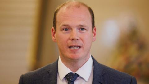 Communities Minister Gordon Lyons during a press conference at Parliament Buildings at Stormont in Belfast - he is wearing a white shirt, grey blazer and grey tie with yellow dots. He has dark hair.
