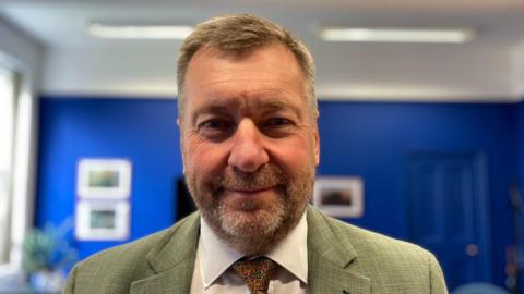 A head and shoulders photograph of Lincolnshire County Council leader Sean Matthews standing in a room with blue walls and a white ceiling. He is wearing a green blazer with a white shirt and paisley tie.