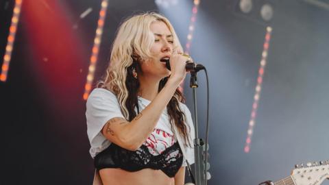 Nieve has blonde and brown hair. She is on a stage, holding a microphone that is on a stand. She is holding a guitar. She is wearing a black bra on top of a white t-shirt