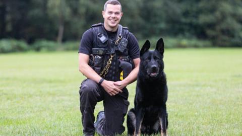 PC Chris Duffy kneels down beside Reno, a black dog who is sitting in a large grassed area with woodland in the background. PC Duffy is smiling at the camera and wearing a black t-shirt and trousers. He is also wearing a black vest with a radio, handcuffs and other police equipment attached to it.