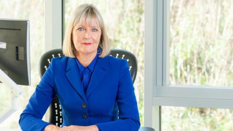 Joy Allen is sitting on a plastic, black chair behind a desk. She has mid-length blonde hair and is wearing a blue suit. There are tall glass windows behind her opening up onto a grassy area.