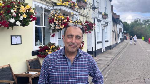 Hossey Saghri is slightly smiling stood outside a yellow inn bedecked in colourful hanging baskets. He has one hand on his hip, receding dark hair with his glasses positioned on the top of his head and a blue checked shirt.