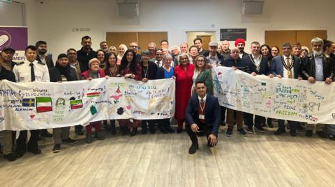 Group of people holding up a long white piece of silk with drawings and words on to describe their feelings on integration and race equity. The banner includes different languages and flags in many colours.