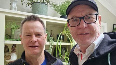 A selfie of Robert and David Taylor standing in front of a kitchen cabinet with leafy green house plants on the shelves. They are wearing outdoor clothes and are smiling.