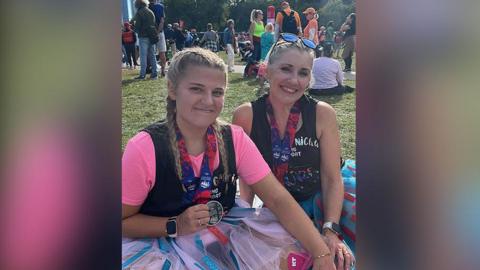 Sitting in a grassy area in Chicago are Gracie Nuttall - who has her long blonde hair in plaits - and her mum Nicola Nuttall, who has her blonde hair tied up. They are wearing marathon medals and are both smiling. Gracie is wearing a bright pink tutu while Nicola's is blue and red.   