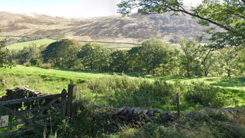 The land surrounding Brockholes Farm which consists of green fields, trees and hills.