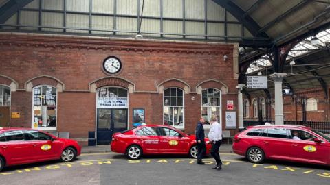 Three red taxis are parked outside of Darlington station at the taxi rank. Two men are stood beside the taxis speaking.