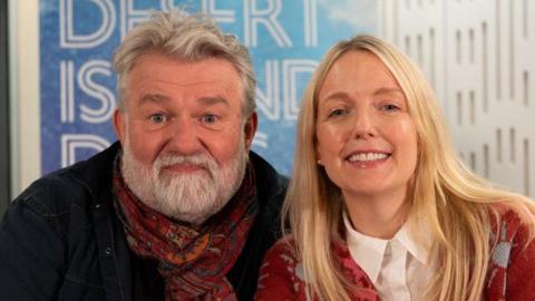 Si King and Lauren Laverne smiling into the camera in front of a blue Desert Island Discs sign. Si is smiling with his lips pursed and has white, short hair and a white beard. He is wearing a black jacket and red paisley scarf which is tied in a knot around his neck. Lauren Laverne is sitting to the right of him (as you look towards them) and has long, blonde hair. She is smiling with her teeth and has glossy lips. She is wearing a red and blue patterned cardigan with a white shirt underneath with is buttoned all the way to the top.
