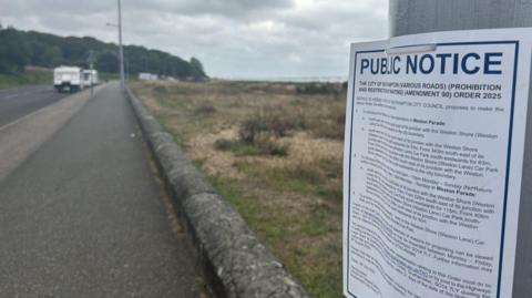 Public notice sign on the right attached to a lamppost with path and road where several caravans are parked in the distance.