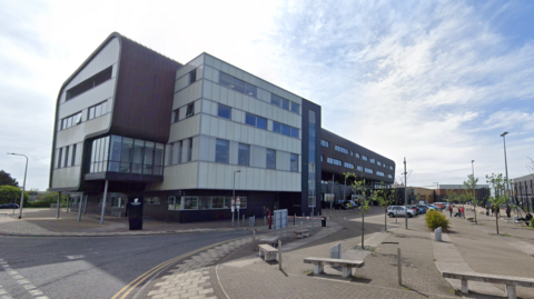 The four-storey Furness College campus building is a mixture of brown and white cube shapes. The front cube juts over the entrance. There are rows of rectangular windows. There are cars parked and benches in the courtyard on the right.