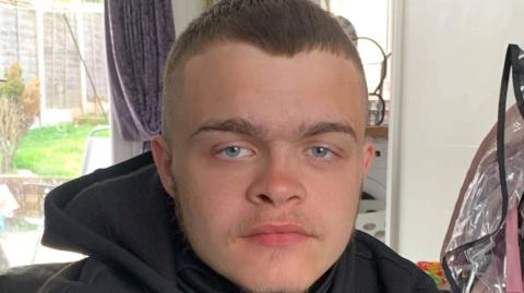 A teenage boy in a black hoodie, sat on a sofa inside a living room. He is looking at the camera.