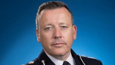 A close-up photo of the acting chief constable of West Midlands Police. He wears a black police uniform and stands in front of a blue background, looking into the camera. 