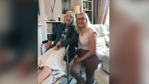 On the left is Irene Wright in a wheelchair - she has grey hair down to her shoulders and glasses, in a green cardigan, a flowery top and a beige skirt.

On the right is Joy Roberts who is kneeling down - she has blonde hair down to her shoulders and glasses, wearing a pink top and brown trousers.