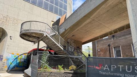 A concrete staircase with metal railings has a twist as it rises to a concrete bridge which is dotted with graffiti. The staircase is cordoned off by metal fencing. There is a tall concrete building, similarly tagged, to the left and an older dark brick building with dark greay windows behind the bridge, of which only the ground floor is visible. On one of the temporary fences, a banner shows the logo of a company that claims to be "Transforming spaces".
