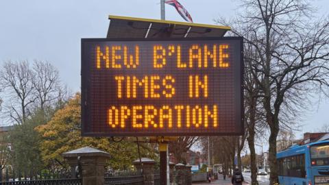 A digital sign that states new b'lane times in operation. Oange and white cones are around the sign. A yellow frame is holding it up. A union flag is in the background. There's a blue bus on the bus lane and cars on the other lanes. It's a wet day with puddles in the street. 