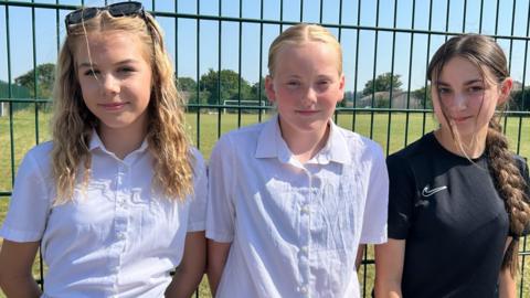 Three young girls, aged around 12-14, standing in a line in front of metal railings with a football pitch in the background. The girl on the left has long dark blonde hair with sunglasses perched on top of her head and is wearing a white blouse. The girl in the middle is also wearing a white blouse and has strawberry blonde hair tied back in a centre parting. The girl on the right has long dark hair in pigtails and is wearing a black Nike t-shirt.