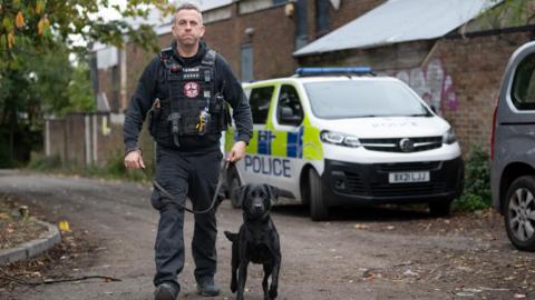 Stuart Philips has short grey hair and is wearing navy blue security-type clothing and a black padded vest. He is holding a lead with a black Labrador-type dog attached. They are walking along a country lane with a police van and large brick building behind them.