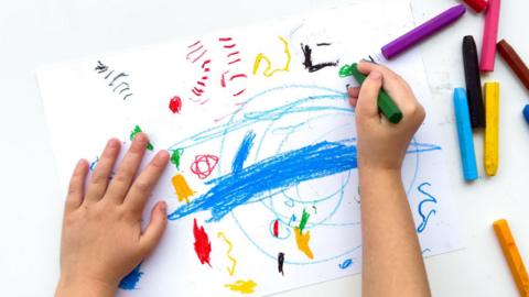 Childs hands drawing with colorful wax crayons on white paper, top view