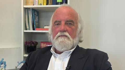 Dai Davies sits in a dark jacket and white shirt with no tie in front of a bookshelf