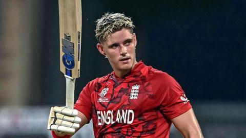 Jacob Bethell, in red team shirt, raises his bat after scoring a century for England
