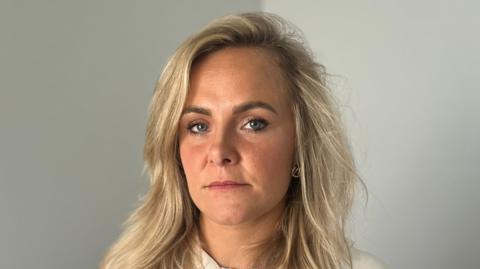 Hannah Farrell, a nurse, looks into the camera. She has a neutral expression on her face. We can see her from the shoulders up. She has shoulder-length blonde hair and is wearing a white or cream top. She has a metal chain around her neck. She is standing in a room, there is a plain grey-coloured wall behind her.