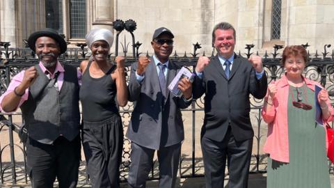 Campaigners pose with raised fists outside a court building after a legal victory. The group stand in front of ornate railings and stone Gothic-style architecture, with one person holding court documents.