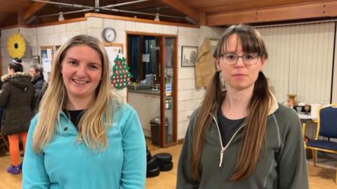 Two women in a community hall smile at the camera. On the left is a woman in a turquoise top, with long blond hair. On the right, a woman with long brown hair in bunches is wearing glasses and a green and beige fleece.There are chairs and tables and a Christmas tree decoration in the background.