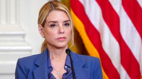 close-up of Pam Bondi, wearing blue suit, looking serious in front of a microphone