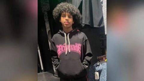 Mohanad Abdullaahi Goobe is photographed with black curly hair. He is pictured wearing a dark hoodie with bright pink writing on it.
