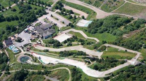 A birds eye view looking down on a dry ski slope snaking through a grass area with trees and buildings