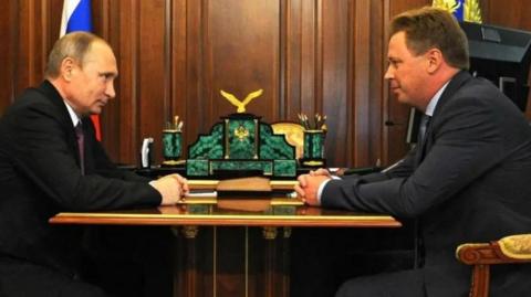 A picture of Vladimir Putin and Dmitrii Ovsiannikov sat opposite eachother. Both men are wearing dark-coloured suits and are sitting at a gold table 