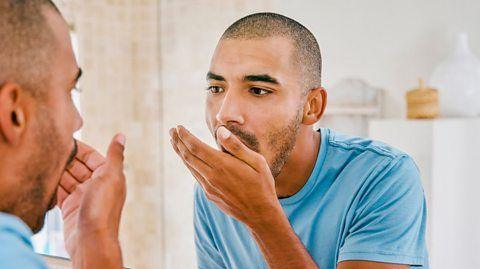 A man checking his breath by holding his hand by his mouth in front of a mirror.