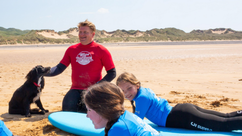 Ariel the dog sitting on a beach while being stroked by a man in a wetsuit with two girls on surfboards