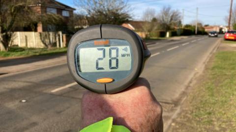 A hand is holding a speed monitoring machine displaying "28mph" on its screen. A road can be seen with a few cars on it. To the left of the road is houses and to the right is grass.