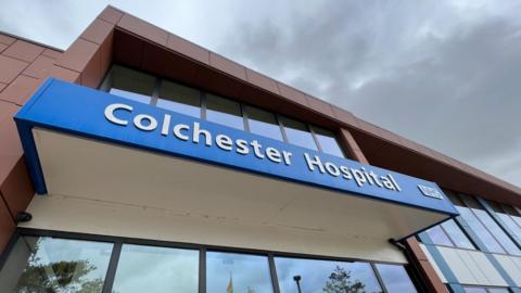 "Colchester Hospital" is written in white font on a blue banner above the hospital's entrance doors. The doors are glass, as is the whole facade.