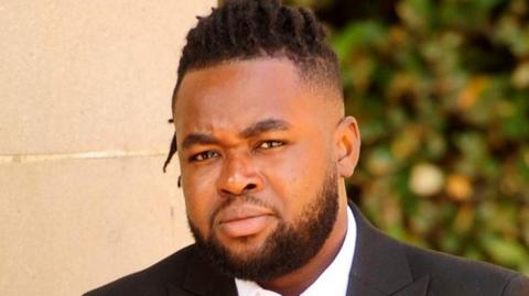A man with a beard and dark hair, closely cropped at the sides, wearing a suit, pictured outside a court