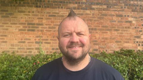 A man stands outside in front of a brick wall. He has a brown hair styled into a short mohawk and a short beard. He is wearing a black t-shirt and a silver chain around his neck, which is tucked under the t-shirt. There is a hedge running along the brick wall behind him.