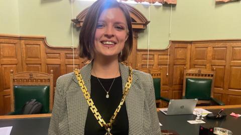 A young woman with shoulder-length brown hair. She has a nose piercing and a lip piercing. She is wearing a thick gold chain over a black and white jacket. 
