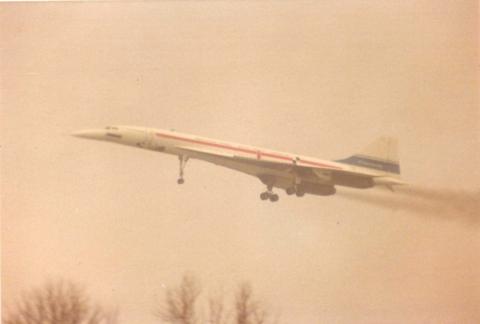 The Concorde prototype is seen taking off into the air. It is white with a red and a blue stripe.