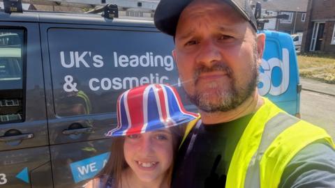 Stuart Field and his daughter take a selfie next to a grey van. He is wearing a high viz jacket and a black cap. His daughter is seen hugging him while wearing a Union Jack bowler hat. 