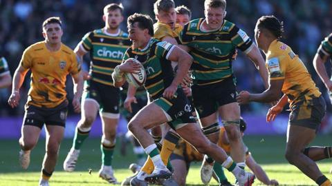 Northampton Saints George Furbank, in a green, yellow and black shirt, runs forward with the ball through a pack of Newcastle Red Bulls defenders.