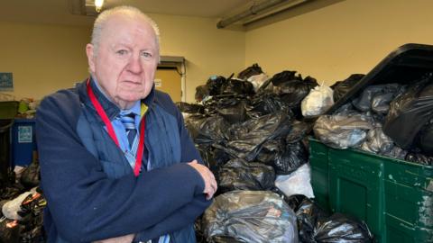 John Harrison standing next to a massive pile of bins. He has his arms crossed. He has thin, greying hair. He is wearing a blue shirt, tie and a blue jacket