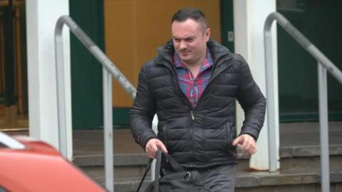 Antony James is leaving a building which has steps and a handrail. His face is scrunched up and he has short dark brown hair. He is wearing a red and blue checked shirt underneath a black puffer jacket and grey jeans. He is holding a black bag. 