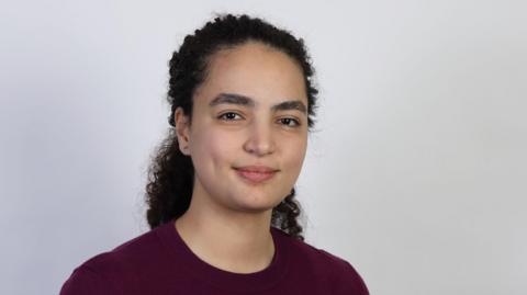 Dr Madjda Bougherira stands in front of a grey background. Her dark curly hair is pulled away from her face. She wears a burgundy jumper