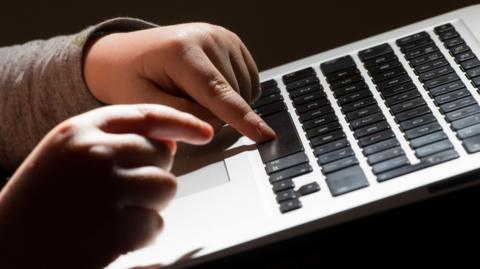 A child's hands typing on a laptop. The laptop's keyboard is black and the child is wearing a long sleeve grey top.