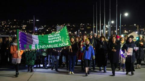 The photo shows people marching down a promenade in Penzance at night. There is a green banner that says "end abuse now".