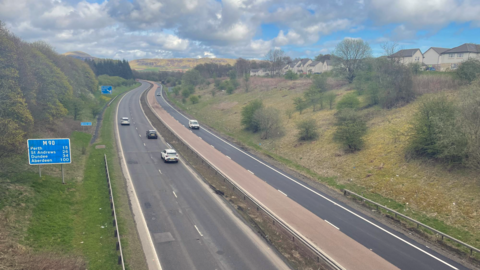M90 with cars on it. There are tyres and houses surrounding it and the sun is shining on hills in the distance.