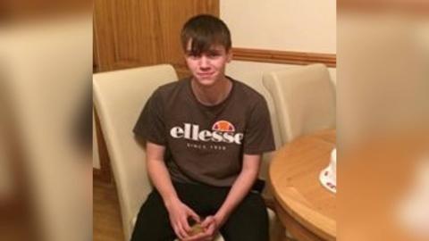 George Joseph Powell sitting on a cream chair, wearing a brown T-shirt and smiling into the camera 